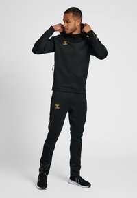 Zwarte sportieve hoodie met een kraag en een geel logo, gecombineerd met zwarte tapervormige broeken met gele accenten en ritszakken.