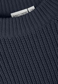 Tessuto di maglione grigio scuro a maglia con colletto a coste. Etichetta visibile con la dicitura "basic needs" posizionata vicino al collo. Motivo testurizzato evidente.