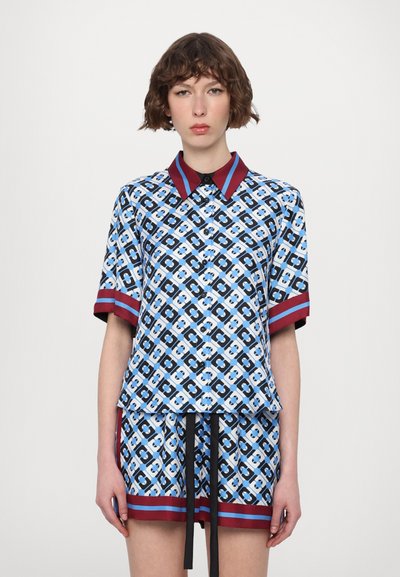 Knop-up korte mouwen top in blauw, zwart en wit met een geometrisch patroon; heeft een rode kraag en bijpassende mouwmanchetten.