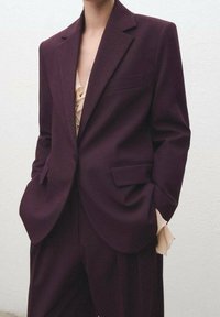 Costume violet foncé avec une veste cintrée et un pantalon à jambes larges, doté d'une poche avant, accompagné d'un haut beige superposé avec des volants.