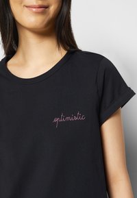 Svart t-shirt med kort ärm, tillverkad av mjukt tyg, med rosa kursiv broderi av ordet "optimistisk" på vänster bröst.