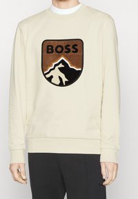 Cream sweatshirt featuring a large embroidered brown and black mountain design with "BOSS" text above. Standard crew neckline and ribbed cuffs.