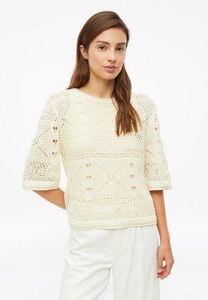 Jonge vrouw met lang bruin haar, draag een crèmekleurige gebreide trui met geometrische patronen en witte broek, staand tegen een effen achtergrond.