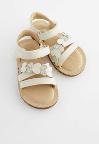 Weiße Sandalen mit einer weichen, strukturierten beigen Innensohle. Sie verfügen über zwei breite Riemen, die mit silberglitzernden Blumenakzenten verziert sind. Flache Sohle.
