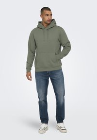Olivengrøn hættebluse med kængurulomme, har ribbede manchetter og kant, parret med blå denimjeans og hvide sneakers.