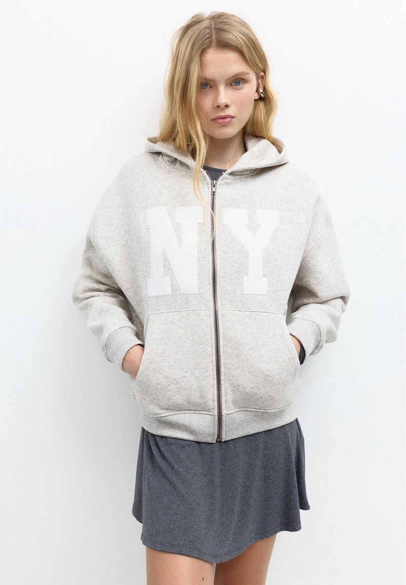 Jeune femme aux cheveux blonds portant un sweat à capuche zippé gris clair avec le texte "NY" et une robe gris foncé sur un fond blanc.