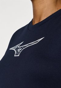 Navy zils T-krekls ar baltu logo, kas veidots ģeometriskā dizainā. Audums šķiet gluds, ar ribinātu apkakles apdari.