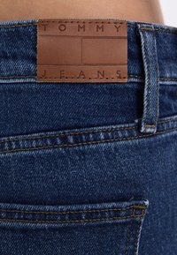 Tommy Jeans mėlynos džinsinės kelnės su rudos odos etiketėmis su įspaustu tekstu, tiesiu kirpimu, matoma siuvinėjimo linija ir galinės kišenės dizainu.