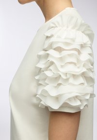 Top bianco in tessuto con maniche a strati ondulate in un materiale trasparente, dalla texture distintiva e un corpo aderente, che mette in risalto una silhouette pulita.