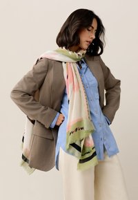 Sciarpa leggera con motivo a fantasie in colori pastello, principalmente rosa e verde, drappeggiata su un blazer marrone e una camicia blu. I bordi sono sfrangiati.