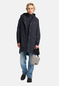 Marineblauwe waterdichte parka met een capuchon, twee voorzakken en een rits. Draag het over een gewatteerde zwarte jas, gecombineerd met lichtblauwe jeans en donkere schoenen.