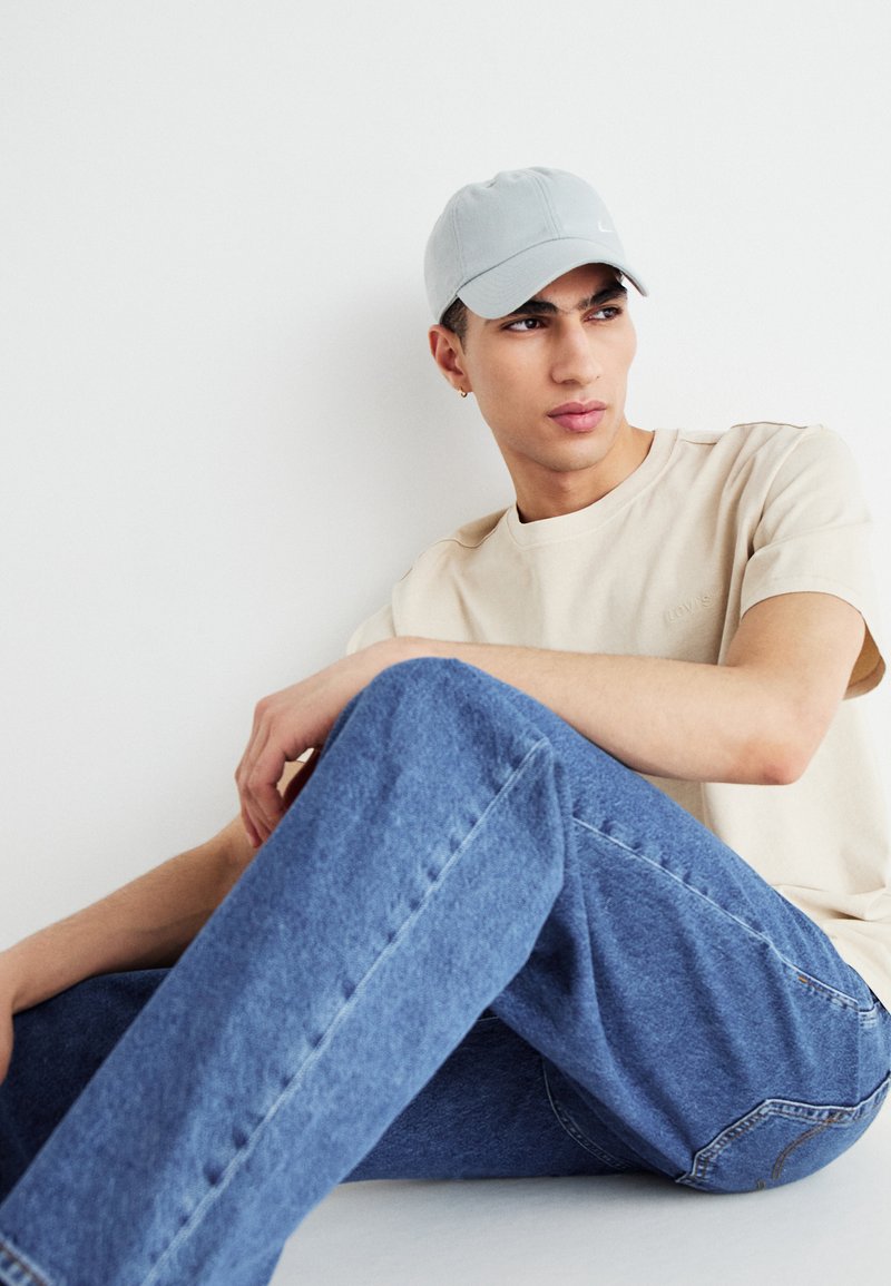 Cappello grigio, T-shirt beige, jeans blu. Posizione seduta, sfondo chiaro, abbigliamento casual, tessuto di media consistenza, branding minimo sulla maglietta.