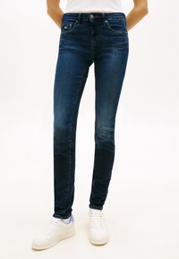 Personne portant un jean slim bleu foncé et des baskets blanches debout sur un fond clair uni.