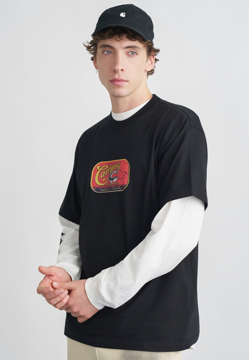 Camiseta negra de manga corta con un gráfico retro en rojo y amarillo. Camiseta blanca de manga larga debajo. Gorra de béisbol negra puesta.