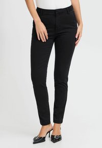 Sieviešu piegulošas melnas bikses, kas izgatavotas no mīksta auduma ar gludu tekstūru, ar klasisku jostu un elegantu siluetu. Apvienotas ar melna krāsas augstpapēžu kurpēm ar smailu purngalu.