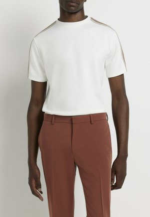 T-shirt blanc à manches courtes avec un col rond et des accents marron contrastants le long des épaules, associé à un pantalon marron ajusté.