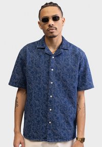 Short-sleeve button-up shirt in dark blue with a textured pattern, featuring a collar and white buttons, paired with sunglasses and a watch.