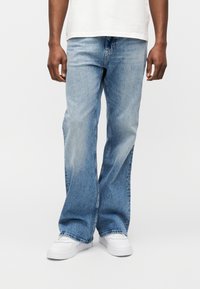 Άνδρας που φοράει ανοιχτό μπλε τζιν παντελόνι κανονικής γραμμής με λευκό μπλουζάκι και λευκά αθλητικά παπούτσια, στέκεται μπροστά σε απλό φόντο.