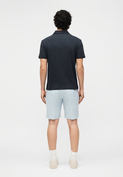 Άνδρας με σγουρά σκουρόχρωμα μαλλιά, φοράει μπλε πόλο μπλούζα, ανοιχτό μπλε σορτς, άσπρες κάλτσες και μπεζ αθλητικά παπούτσια, στέκεται γυρισμένος με την πλάτη.