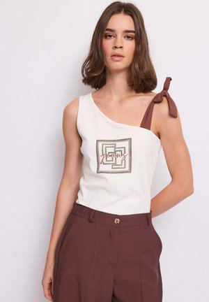 Giovane donna con capelli castani di media lunghezza indossa un top bianco asimmetrico con fiocco marrone e pantaloni marroni a vita alta su sfondo neutro.