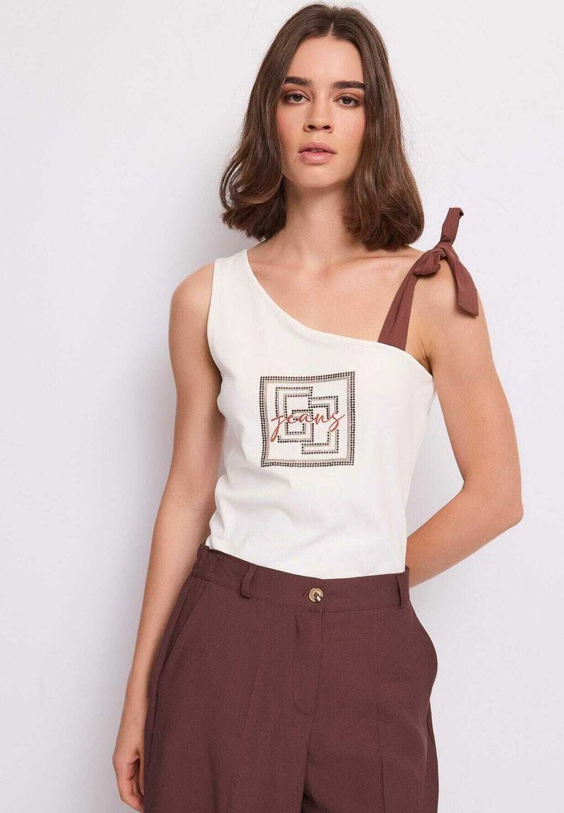 Giovane donna con capelli castani di media lunghezza indossa un top bianco asimmetrico con fiocco marrone e pantaloni marroni a vita alta su sfondo neutro.