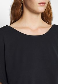 Černý top s krátkým rukávem a širokým, zaobleným výstřihem. Vyrobeno z hladkého materiálu, s volným střihem a minimalistickým designem.