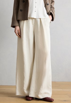 Γυναίκα με κρεμ παντελόνι φαρδύ στα πόδια, λευκό πουκάμισο με κουμπιά, καφέ σακάκι και βυσσινιά παπούτσια, που στέκεται σε ξύλινο πάτωμα.