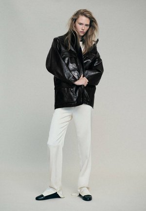 Jeune personne aux longs cheveux porte une veste en cuir noire oversize, un pantalon blanc et des chaussures noires, debout devant un fond clair uni.