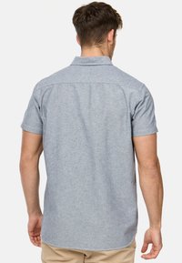 Camisa de manga corta, con cuello, en tela azul clara con un patrón texturizado. Presenta un dobladillo curvado y costuras sutiles a lo largo de los hombros.