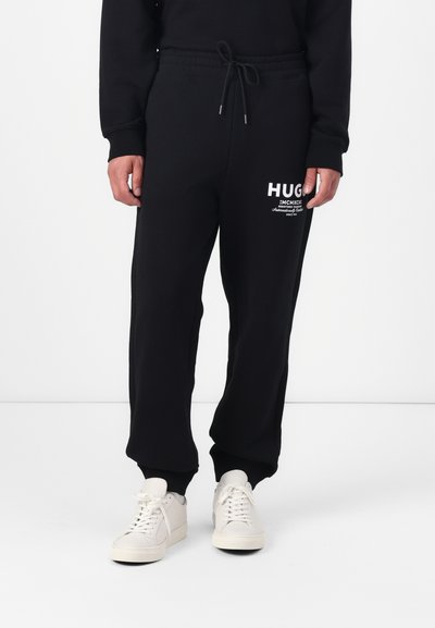 Pantalons de survêtement noirs avec une taille élastique et un cordon de serrage. Présente le logo HUGO blanc imprimé sur le devant. Associés à des baskets blanches.