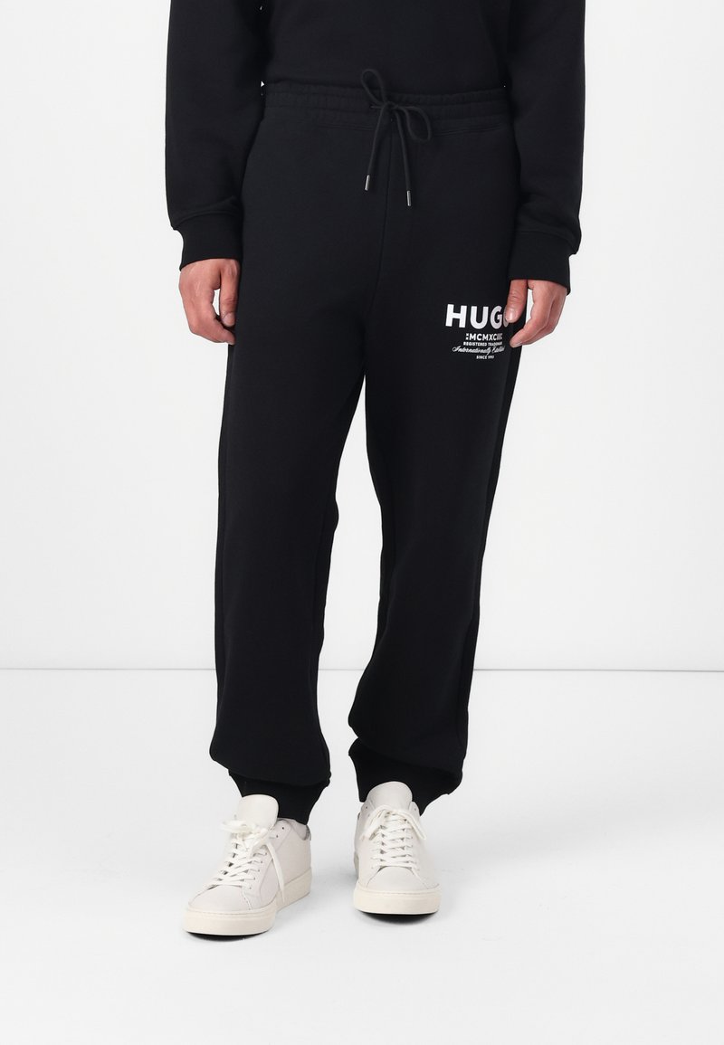 Black sweatpants with an elastic waistband and drawstring. Features white HUGO logo printed on the front. Paired with white sneakers.