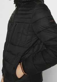 Czarna kurtka puchowa z poziomymi pikowanymi sekcjami, zapinana na zamek błyskawiczny, z kapturem. Posiada pomarańczowe akcenty na mankietach oraz małą naszywkę z logo.