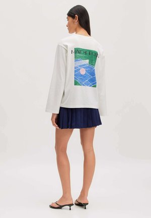 Vrouw met lang donker haar die een wit shirt met lange mouwen draagt met een tennisveldafbeelding en een marineblauwe geplooide rok, van achteren gezien.