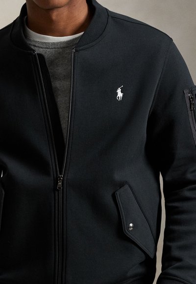 Mees kannab musta lukuga jopet, millel on valge polo mängija logo rinnal, klapiga tasku suru-klõpsuga ja lukuga tasku varrukal.