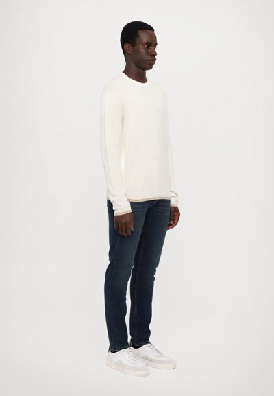 Weißer, gerippter Langarmpullover, dunkelblaue Jeans und weiße Sneakers. Vor einem schlichten Hintergrund. Keine sichtbaren Muster oder Hardware.