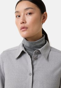 Junge Frau mit dunklen, zurückgebundenen Haaren, die ein hellgraues, knöpfbares Hemd über einem grauen Rollkragenpullover trägt, vor einem schlichten Hintergrund.