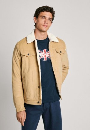 Chaqueta de pana color beige con un cuello de borreguito blanco, dos bolsillos en el pecho y cierres de botón, llevada sobre una camiseta azul marino con un diseño de la bandera británica.