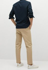 Uomo che indossa una camicia blu navy arrotolata, pantaloni chinos beige e sneakers bianche, in piedi con le mani nelle tasche contro uno sfondo bianco semplice.