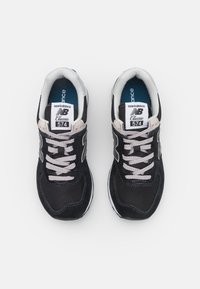 Des baskets New Balance 574 noires présentent une combinaison de matériaux en mesh et en suède, avec des lacets et des accents gris clair, ainsi qu'un col rembourré.