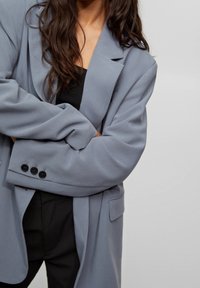 Blazer gris à coupe ample, avec des détails de boutons sur les poignets et une poche avant. Porté sur un haut noir, texture de tissu doux.