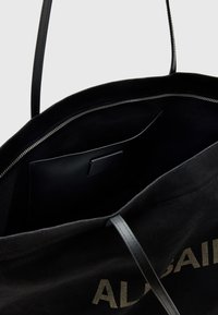 Borsa a spalla in tela nera con manici in pelle, dotata di chiusura con zip e una tasca interna in pelle nera. Scritta dorata sulla parte esterna.