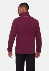 Giacca in pile viola con colletto alto, realizzata in un tessuto tattile e con un logo sulla parte superiore della schiena. Cuciture visibili e posizionamento delle mani.