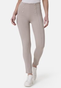 Beige hochtaillierte Leggings mit zwei seitlichen Knöpfen, glatter Textur, detaillierter Naht und taillierter Silhouette. Getragen mit weißen Sneakers.