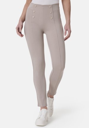 Beige hochtaillierte Leggings mit zwei seitlichen Knöpfen, glatter Textur, detaillierter Naht und taillierter Silhouette. Getragen mit weißen Sneakers.