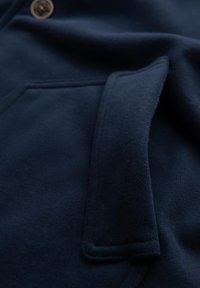 Granatowy sweatshirt o miękkiej fakturze, z kieszenią z przodu i detalem w postaci guzika. Materiał jest gładki o matowym wykończeniu.