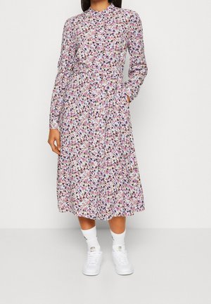 Kobieta ubrana w midi sukienkę w kwiaty z długim rękawem i wysokim kołnierzem, w białych skarpetkach i białych trampkach, stojąca na jednolitym tle.