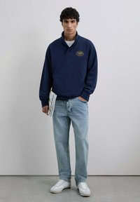 Suéter azul marino con cuello alto y bolsillo frontal, con detalle de logotipo en texto. Pantalones vaqueros rectos de un azul claro y zapatillas blancas.