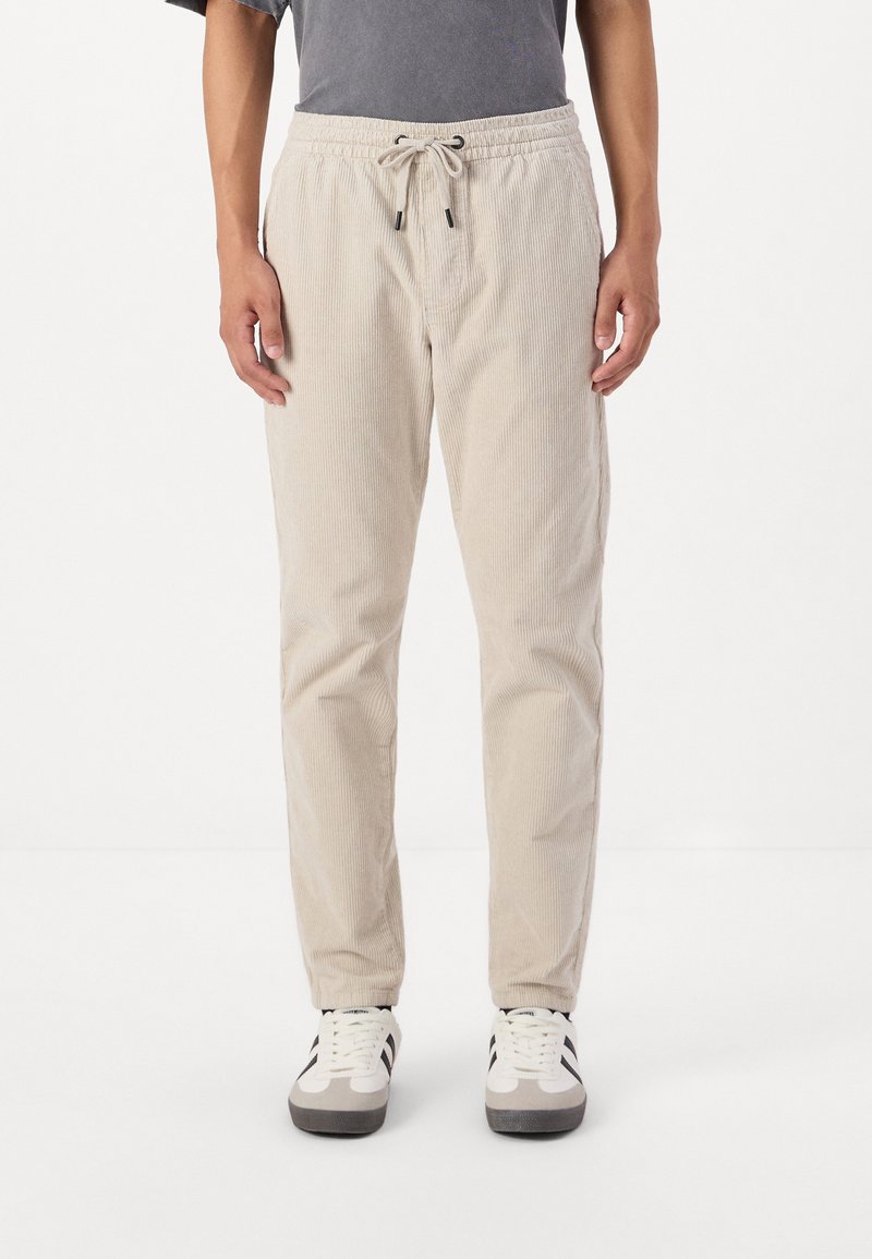Beige Cord-Hosen mit elastischem Bund und Kordelzug, gerade geschnitten und mit seitlichen Taschen, kombiniert mit schwarz-weißen Sneakern.