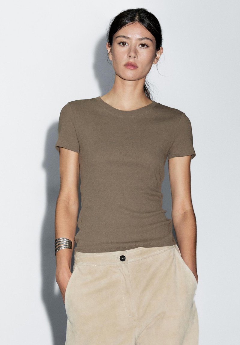 T-shirt marrone aderente con maniche corte, collo rotondo e texture liscia, abbinato a pantaloni beige dotati di tasca frontale e bottone.