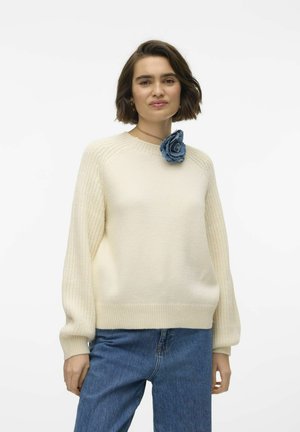 Maglione lavorato a maglia color crema con maniche a coste e collo rotondo, decorato con una rosa di tessuto blu sulla spalla. Indossato con jeans blu.